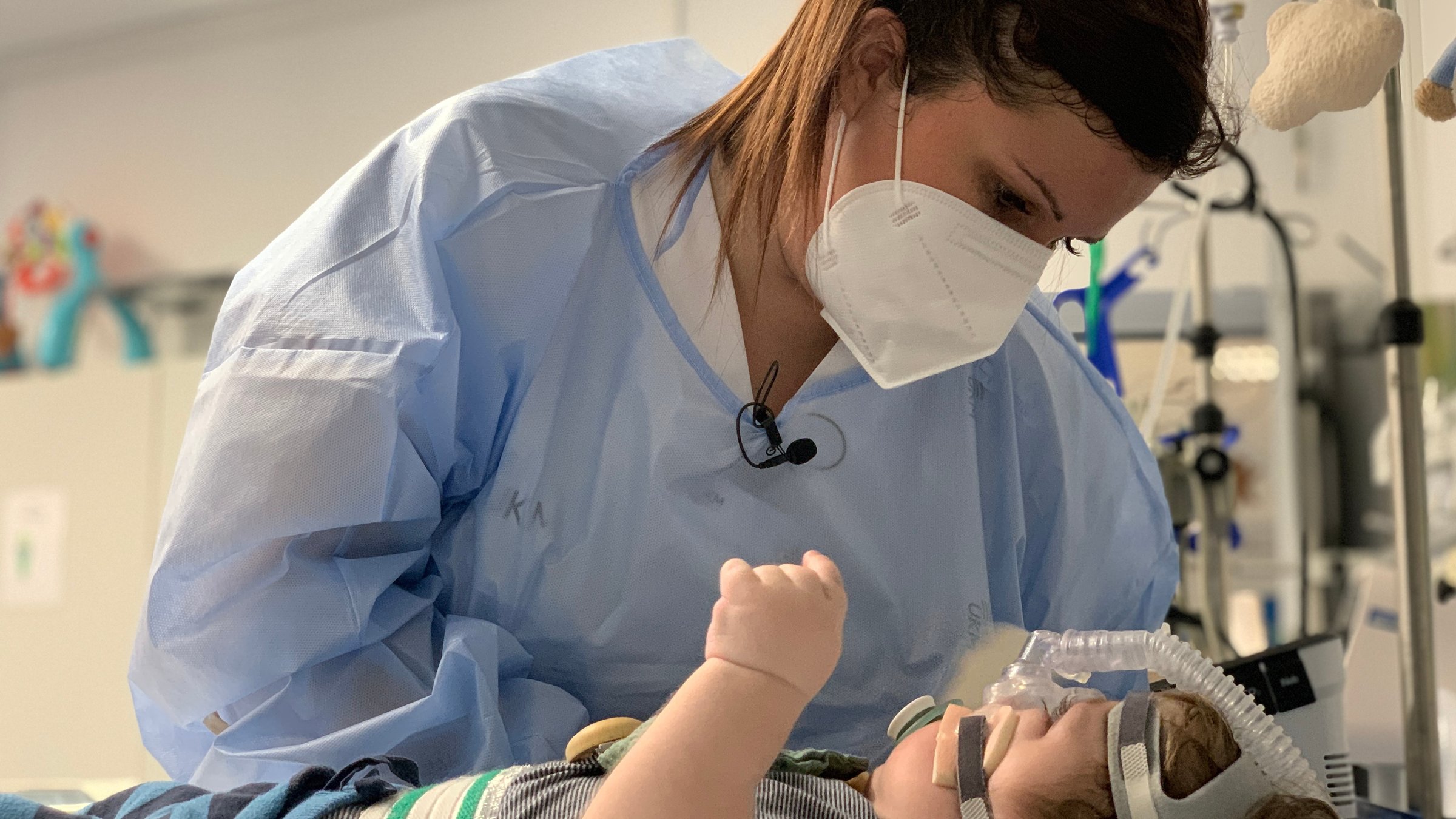 "37°: Notfall Kinderklinik": Eine Ärztin mit dunkelbraunen Haaren, die zu einem Zopf hochgebunden gebunden sind, trägt eine weiße FFP2-Maske und einen blauen Schutzmantel über ihren weißen Kittel. Sie beugt sich über ein Kleinkind, dass auf der Trage liegt und ein Beatmungsgerät am am Kopf befestigt hat.