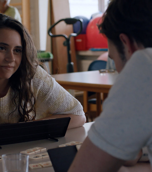 "Sense of Tumour - Tsunami": Tristan (Maarten Nulens) und Hanne (Marthe Schneider) sitzen sich in einem Aufenthaltsraum gegenüber und spielen etwas. Dabei schaut Hanne Tristan aufmerksam an.