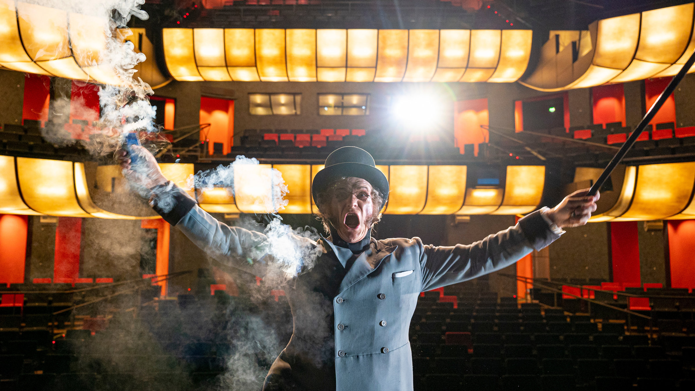 "Beethoven - Ein Geisterspiel": Ein Mann mit grauem Frack und Zylinder steht auf einer Theaterbühne. Hinter ihm sieht man den beleuchteten, aber leeren Theatersaal mit roten Stühlen. In seiner linken Hand hält er einen Stock, aus seiner rechten Hand dampft es. Er hat den Mund geöffnet, so, als würde er rufen oder schreien.