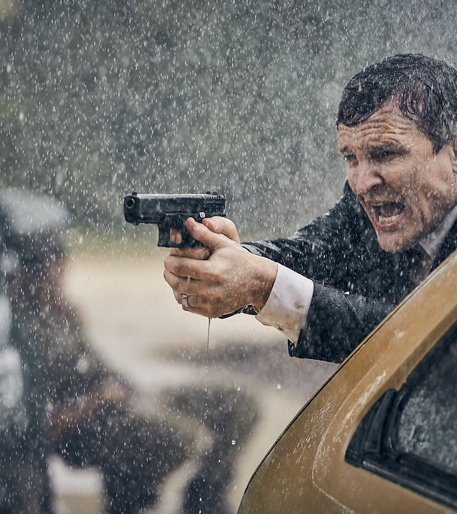 "The Tourist: Hüte dich vor dem Zorn des sanftmütigen Mannes": Detective Inspector Lachlan Rogers (Damon Herriman) steht im strömenden Regen mit gezogener Waffe hinter einem Wagen.