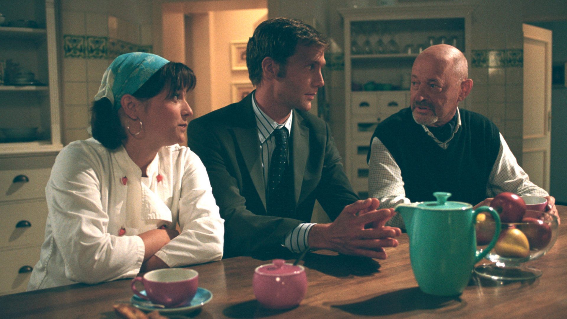 "Bianca - Wege zum Glück: Folge 20": Abends in der Küche: Oliver (Patrik Fichte), Köchin Bärbel (Birgit Wiedel Weidinger) und der Verwalter Georg (Uwe Zerbe) sitzen am, mit bunten Tassen und einer Kanne gedeckten, Küchentisch. Oliver sitzt in der Mitte und redet mit Georg. Bärbel wirkt amüsiert und blickt ebenfalls zu Georg hin.