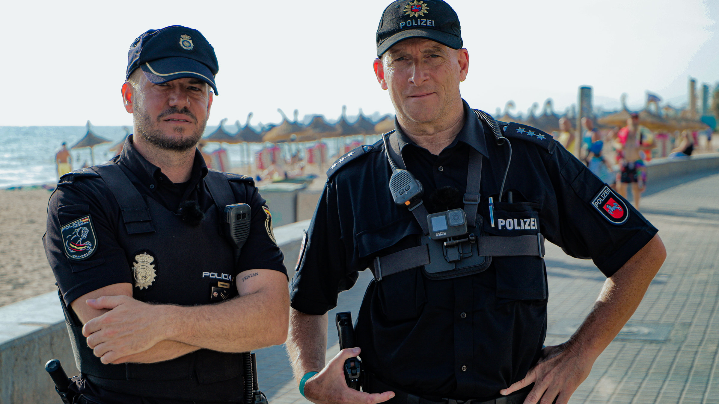 "ZDF.reportage: Streife auf Mallorca - Zoff am Ballermann": Zwei Polizisten, Javier Pulpillo-Mas und Torsten Obst, stehen an der Strandpromenade von Palma de Mallorca. Im Hintergrund liegt das Meer und der Strand, an dem sich Badende befinden.