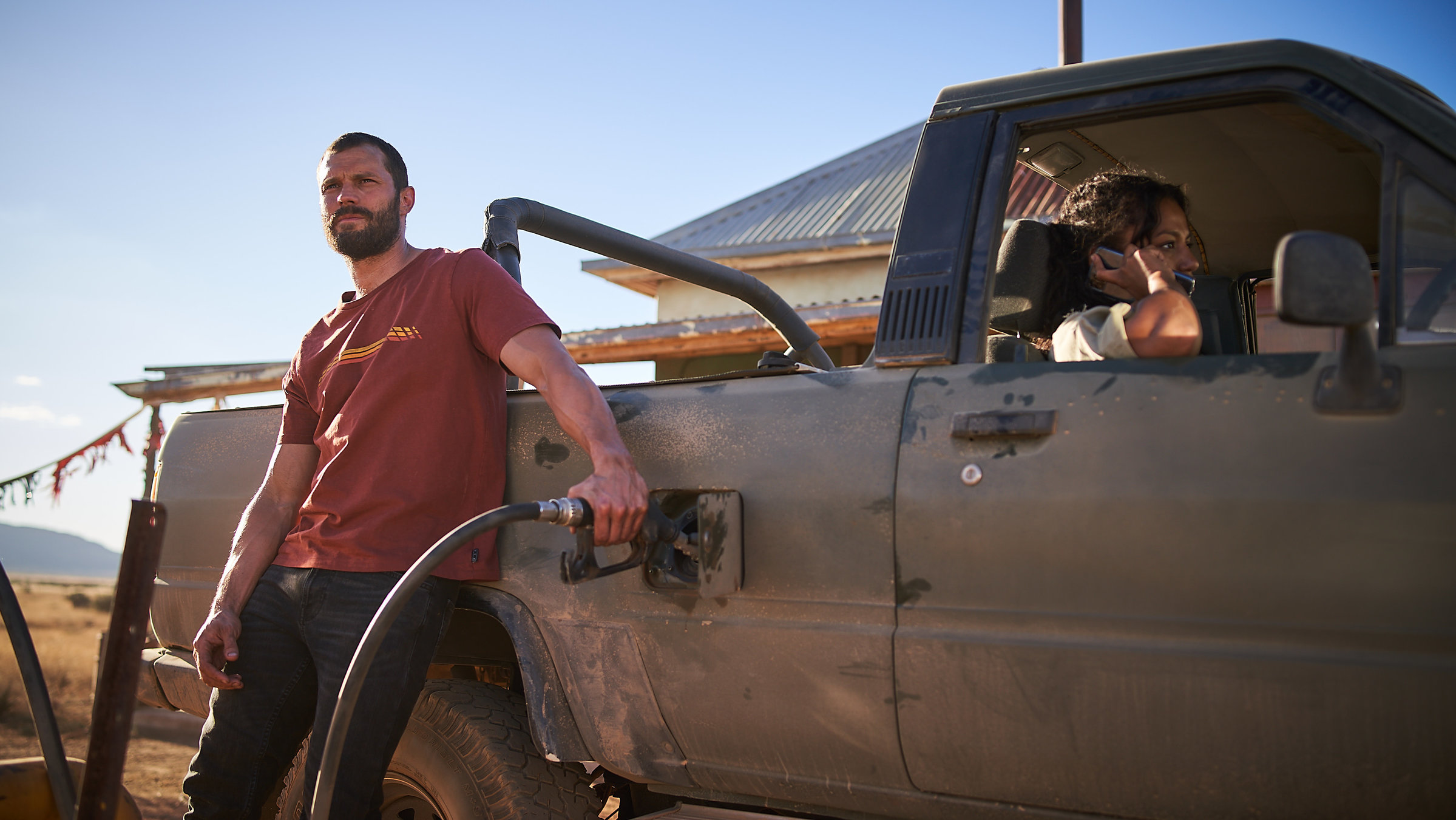 "The Tourist - Duell im Outback: Waiting for the Sun": Der Mann (Jamie Dornan) lehnt lässig an seinem Pick-up und hält den Zapfhahn in den Tank. Luci (Shalom Brune-Franklin) sitzt auf dem Beifahrersitz und telefoniert.