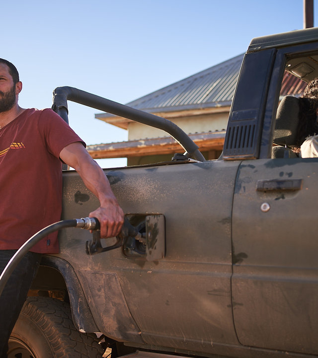 "The Tourist - Duell im Outback: Waiting for the Sun": Der Mann (Jamie Dornan) lehnt lässig an seinem Pick-up und hält den Zapfhahn in den Tank. Luci (Shalom Brune-Franklin) sitzt auf dem Beifahrersitz und telefoniert.