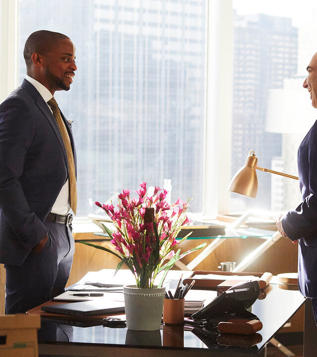 "Suits - Argwohn": Alex (Dulé Hill, l.) und Louis (Rick Hoffmann) stehen sich gegenüber. Zwischen ihnen ein Schreibtisch, auf dem rosafarbene Blumen stehen. Im Hintergrund sind durch das Fenster die Hochhäuser New Yorks zu sehen.
