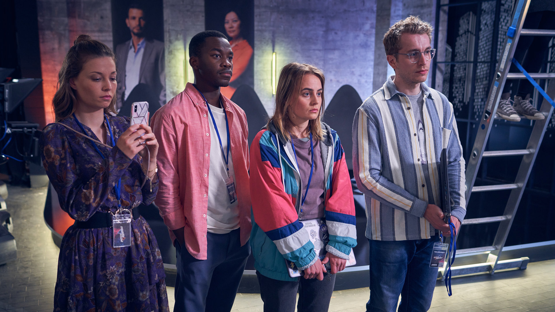 "Start the fck up - Pitch Perfect": Melina (Lena Meckel) blickt auf ihr Smartphone, dass sie in der Hand hält, neben ihr stehen nebeneinander Kenny (Kelvin Kilonzo), Jana (Olga von Luckwald) und Jens (László Branko Breiding) und blicken in die Ferne. Im Hintergrund sind eine Leiter und Studiokulissen zu sehen.