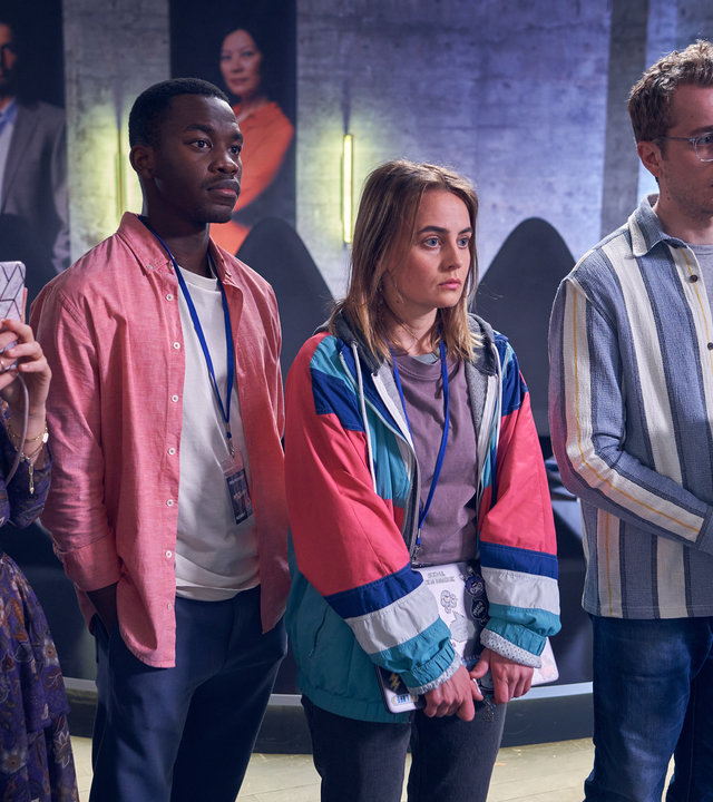 "Start the fck up - Pitch Perfect": Melina (Lena Meckel) blickt auf ihr Smartphone, dass sie in der Hand hält, neben ihr stehen nebeneinander Kenny (Kelvin Kilonzo), Jana (Olga von Luckwald) und Jens (László Branko Breiding) und blicken in die Ferne. Im Hintergrund sind eine Leiter und Studiokulissen zu sehen.