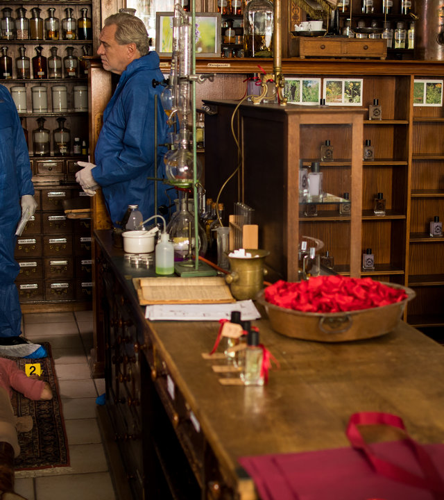 "SOKO München - Duftspuren": In der Parfümerie liegt eine Leiche hinter der Theke auf dem Boden, am Kopf stehen Dr. Weissenböck (Florian Odendahl), der hinter Franz Ainfachnur (Christofer v. Beau) steht und ihn demonstrativ am Kragen packt, gegenüber steht Arthur Bauer (Gerd Silberbauer). Auf der anderen Seite des Ladens sitzt Billie Curio (Sina Reiß) in der Hocke und schaut zum Schrank hinter der Theke.