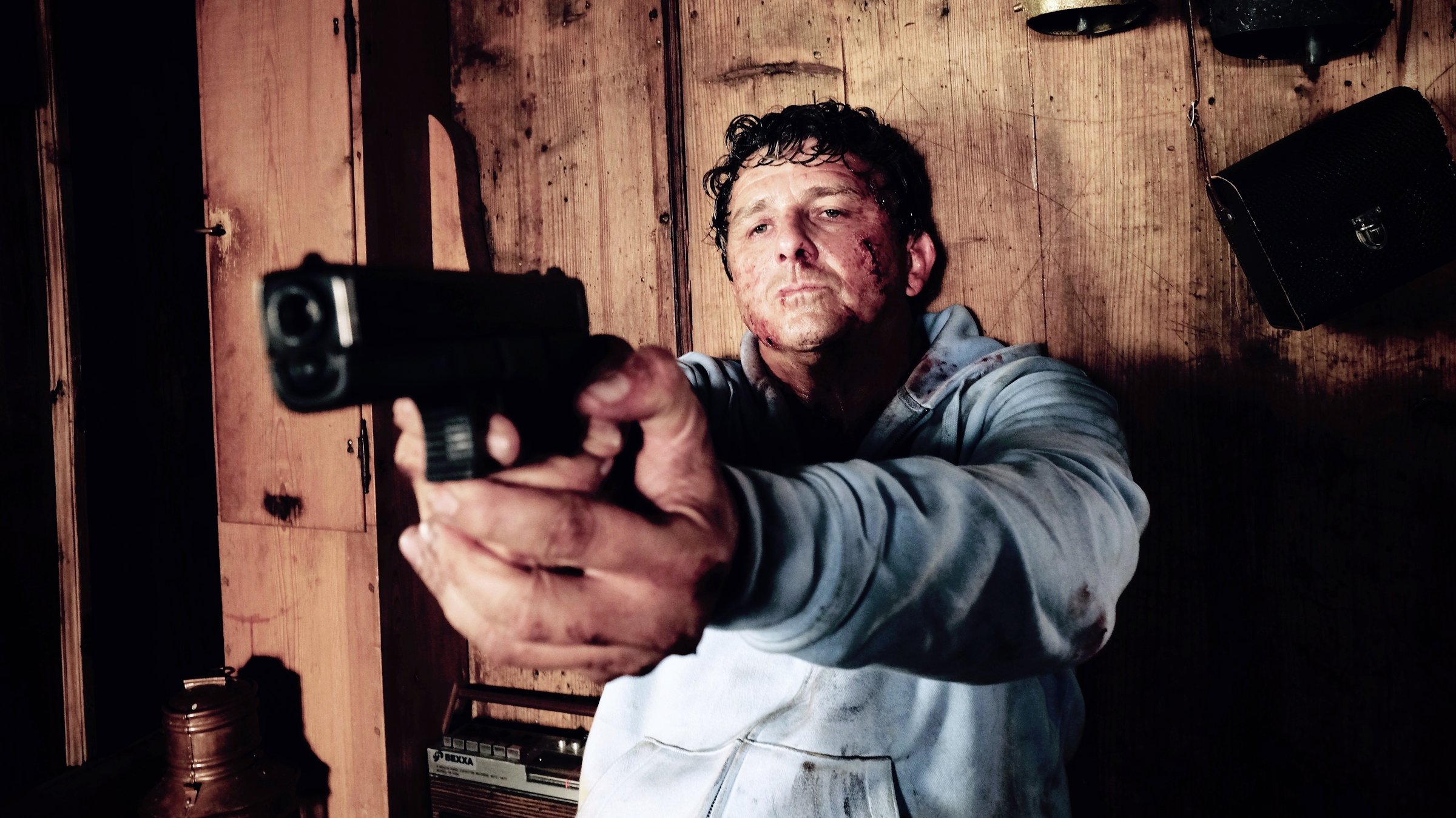 "Flucht durchs Höllental": Rechtsanwalt Klaus Burg (Hans Sigl) sitzt verletzt, mit gezogener Waffe vor einer Holzbretterwand, an der Kuhglocken befestigt sind und eine Umhängetasche hängt. Neben ihm auf der Bank steht ein Kassettenrekorder und in der Ecke eine Milchkanne.