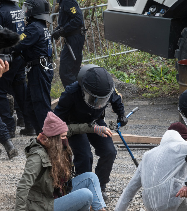 "SOKO Stuttgart – Blutiger Protest": Streikende Mitglieder einer Umweltschutzgruppe (Komparsen) werden von Polizeibeamten in voller Schutzmontur weggezerrt. Einer der Beamten hält eine große Zange in der Hand, um Eisenketten zu lösen. Im Vordergrund sieht man einen Reporter, der die Ereignisse filmt.