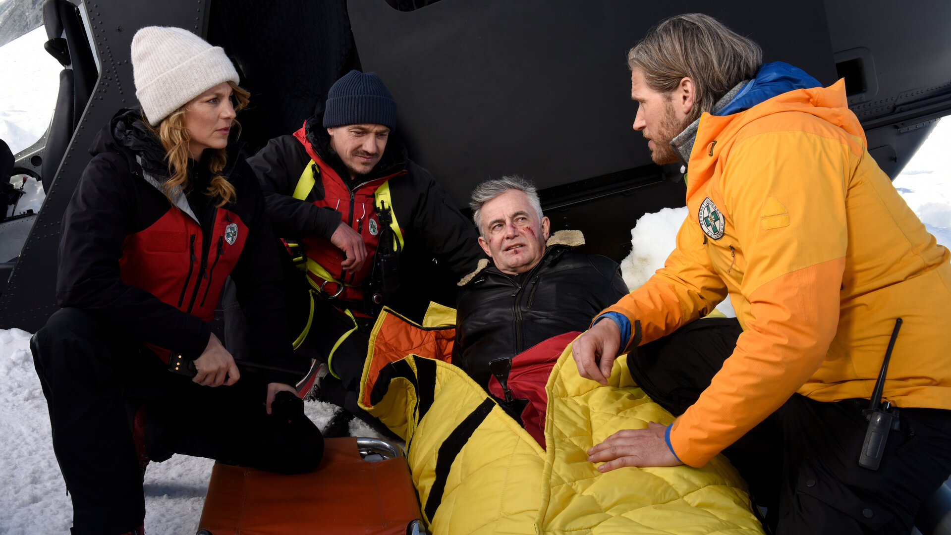 "Die Bergretter - Dünnes Eis": In einer Schneekulisse: Vor dem abgestürzten Hubschrauber liegt Michi (Robert Lohr), mit Verletzungen im Gesicht, eingepackt in eine Rettungsdecke, im Schnee. Er blickt zu Bergretter Markus (Sebastian Ströbel), der neben ihm kniet. Ihm gegenüber sein Bergretter-Team Katharina (Luise Bähr) und Tobi (Markus Brandl). Die drei blicken sich ernst an.