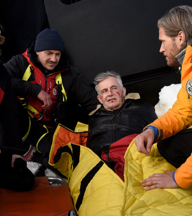 "Die Bergretter - Dünnes Eis": In einer Schneekulisse: Vor dem abgestürzten Hubschrauber liegt Michi (Robert Lohr), mit Verletzungen im Gesicht, eingepackt in eine Rettungsdecke, im Schnee. Er blickt zu Bergretter Markus (Sebastian Ströbel), der neben ihm kniet. Ihm gegenüber sein Bergretter-Team Katharina (Luise Bähr) und Tobi (Markus Brandl). Die drei blicken sich ernst an.
