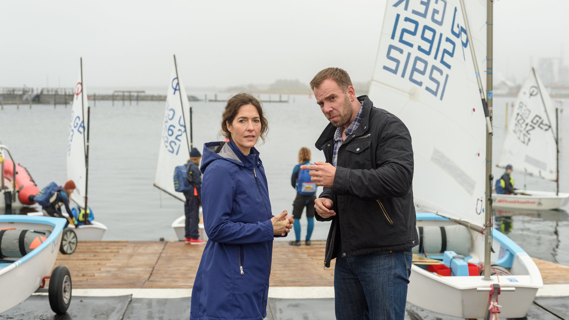 "SOKO Wismar – Außer Atem": Lars Pöhlmann (Dominic Boeer) und Isabell Faude (Margrit Sartorius) stehen sich an der Slipanlage des Segelclubs zwischen einigen Booten im Gespräch gegenüber. Bei diesigem Wetter lassen einige andere Segler im Hintergrund ihre Boote zu Wasser.