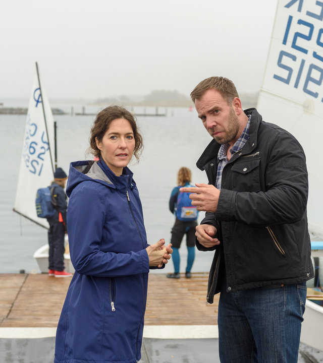 "SOKO Wismar – Außer Atem": Lars Pöhlmann (Dominic Boeer) und Isabell Faude (Margrit Sartorius) stehen sich an der Slipanlage des Segelclubs zwischen einigen Booten im Gespräch gegenüber. Bei diesigem Wetter lassen einige andere Segler im Hintergrund ihre Boote zu Wasser.