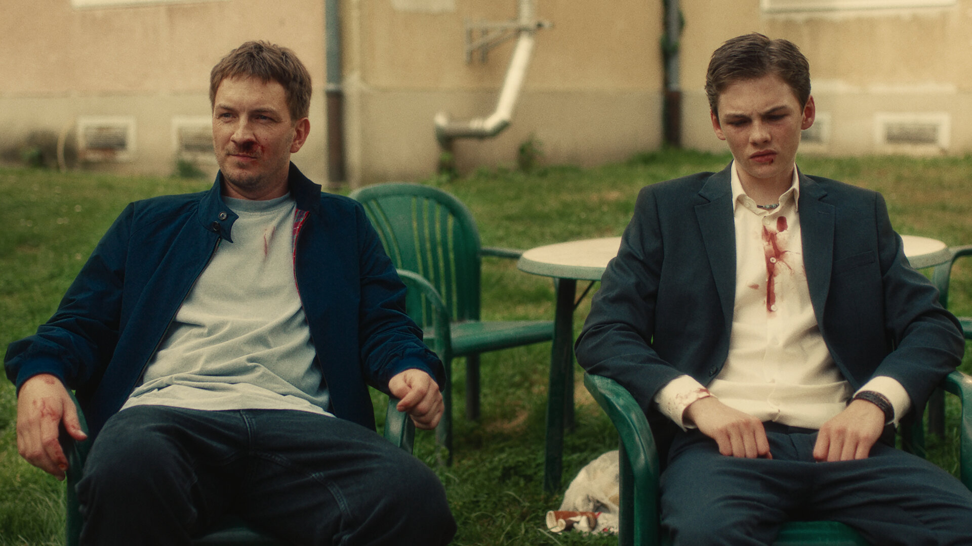 "Chabos - Abschlussklasse 06": Der erwachsene Peppi (Johannes Kienast) und der junge Peppi (Nico Marischka) sitzen erschöpft und beide mit blutiger Nase nebeneinander auf Gartenstühlen vor einem Haus.