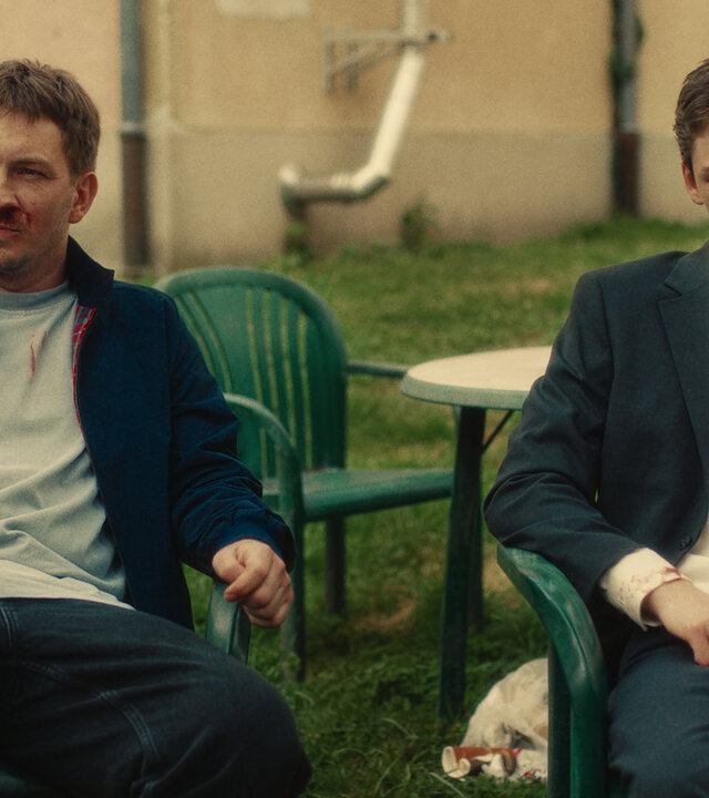 "Chabos - Abschlussklasse 06": Der erwachsene Peppi (Johannes Kienast) und der junge Peppi (Nico Marischka) sitzen erschöpft und beide mit blutiger Nase nebeneinander auf Gartenstühlen vor einem Haus.