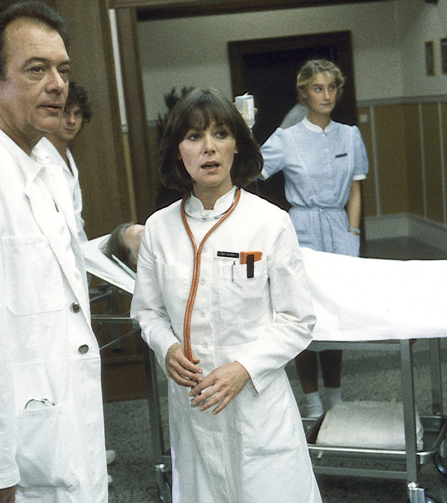 "Die Schwarzwaldklinik - Die Entführung": Professor Brinkmann (Klausjürgen Wussow) und Dr. Elena Bach (Heidelinde Weis) stehen nebeneinander im Krankenhausflur und unterhalten sich. Im Hintergrund sind ein Patient auf einer Liege und mehrere Schwestern zu sehen.