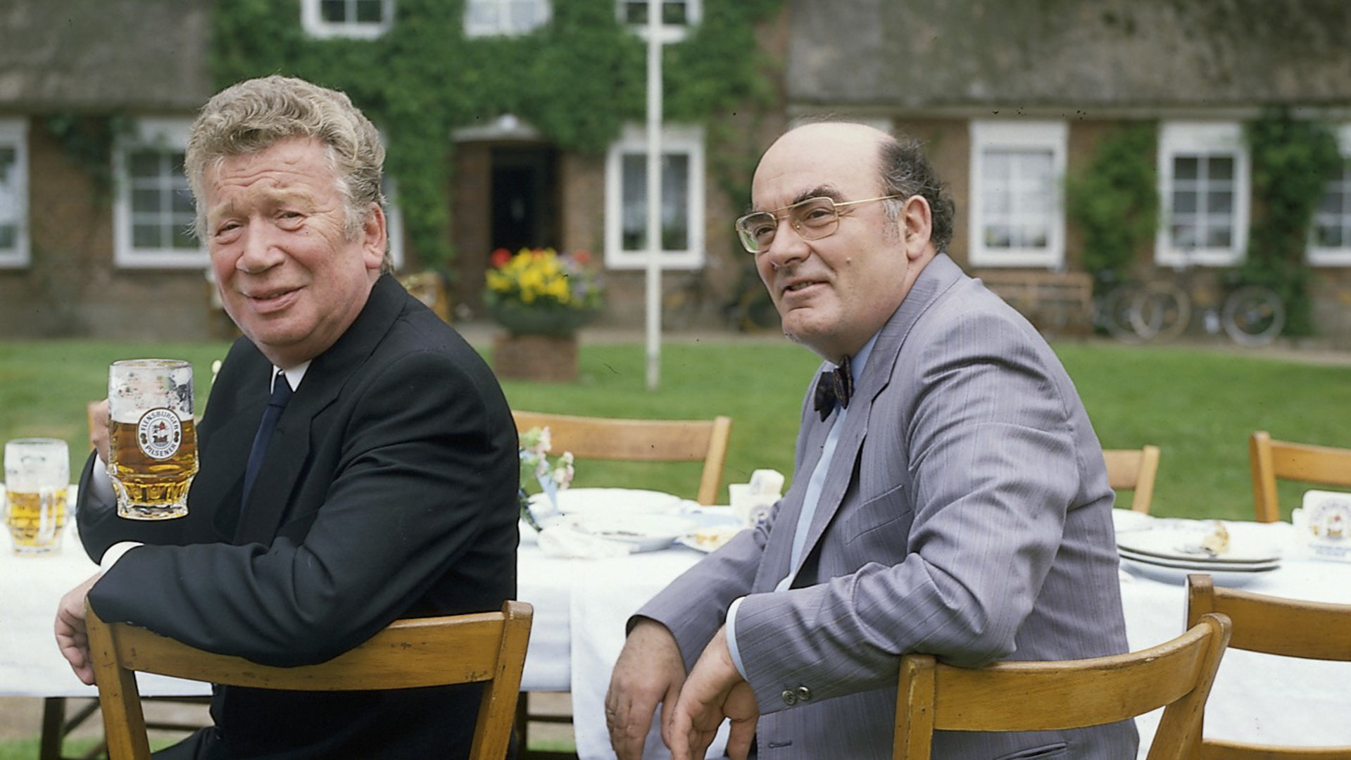 "Der Landarzt - Nachwuchs": Pfarrer Eckholm (Heinz Reincke) und Bürgermeister Schultes (Hans Häckermann) sitzen gemeinsam an einer Festtafel. Der Pfarrer hält ein Glas Bier in der Hand.