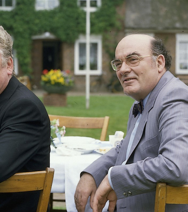 "Der Landarzt - Nachwuchs": Pfarrer Eckholm (Heinz Reincke) und Bürgermeister Schultes (Hans Häckermann) sitzen gemeinsam an einer Festtafel. Der Pfarrer hält ein Glas Bier in der Hand.