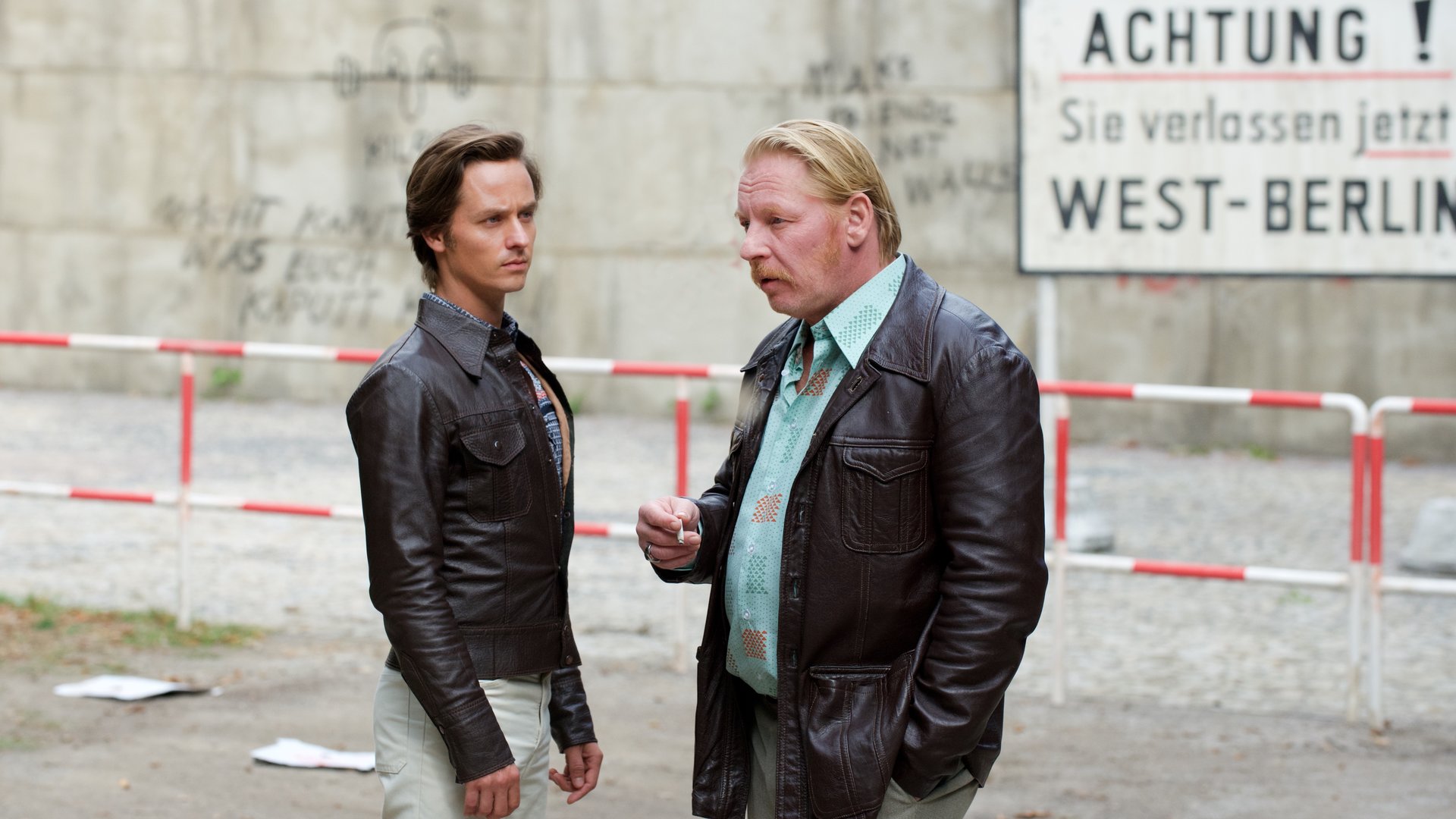 "Der gleiche Himmel (2)": Lars Weber (Tom Schilling) und Ralf Müller (Ben Becker) stehen unweit vom Sperrbezirk und unterhalten sich. Im Hintergrund ist die Berliner Mauer und ein Schild mit der Aufschrift "Achtung! Sie verlassen jetzt West-Berlin" zu sehen.