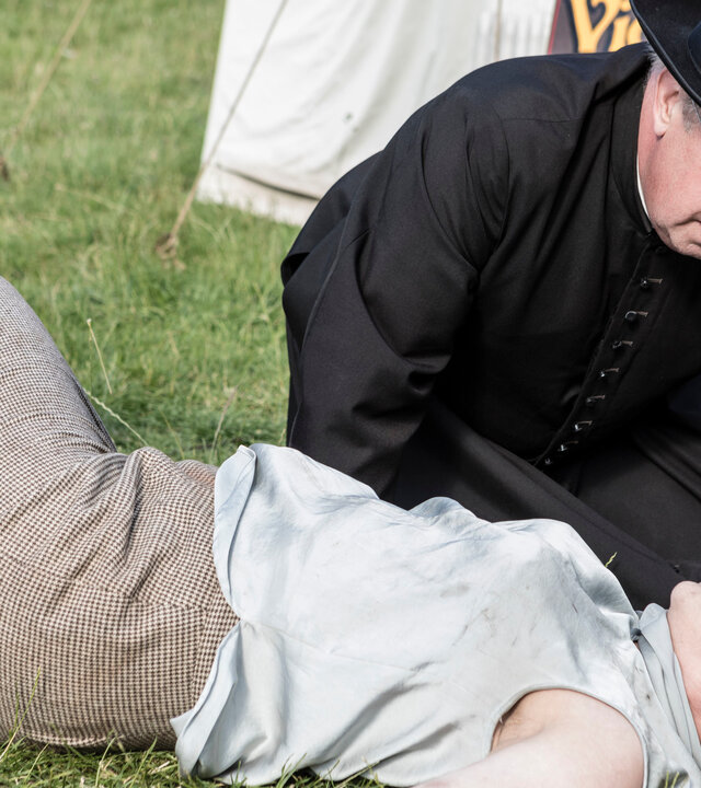"Father Brown - Die Weisheit der Narren": Father Brown (Mark Williams) kniet neben Dr. Belmont (Annabel Cleare), die vor einem der Festzelte zusammengebrochen ist.