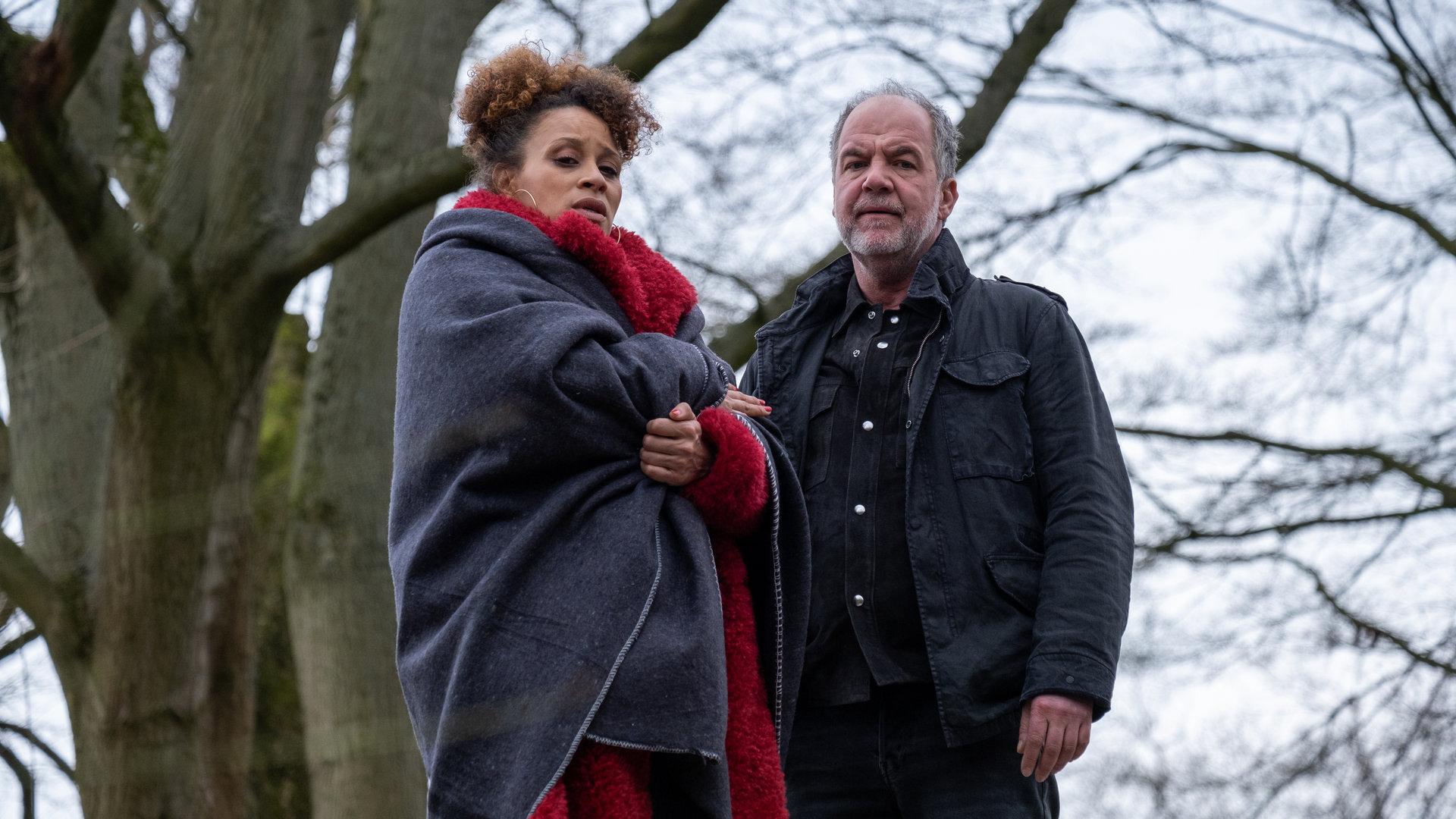 "SOKO Hamburg - Offene Rechnungen": Ella Hansen (Oceana Mahlmann) und Oskar Schütz (Marek Erhardt) stehen nebeneinander im Freien und blicken von oben in die Kamera. Ella hat sich eine Decke umgelegt. Im Hintergrund steht ein großer Baum mit kahlen Ästen.