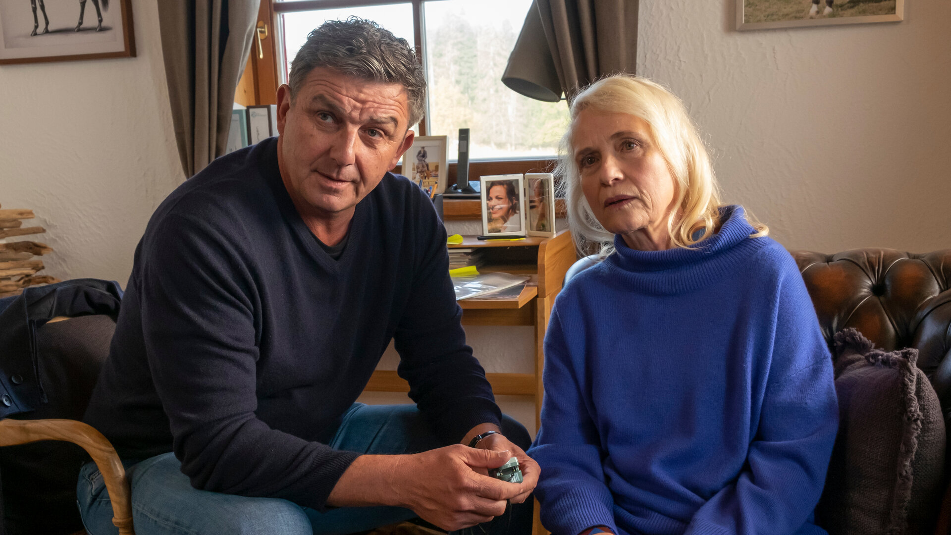 "Der Bergdoktor: Herzschmerzen (2)": Im Wohnhaus der Familie Achinger: Blass sitzt Helen Aichinger (Alexandra von Schwerin) auf einem Sofa. Neben ihr sitzt Dr. Martin Gruber (Hans Sigl), die Stirn nachdenklich in Falten gelegt, auf einem Stuhl und hält einen Finger-Pulsoximeter in der Hand.