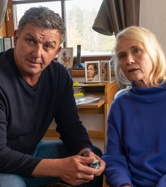 "Der Bergdoktor: Herzschmerzen (2)": Im Wohnhaus der Familie Achinger: Blass sitzt Helen Aichinger (Alexandra von Schwerin) auf einem Sofa. Neben ihr sitzt Dr. Martin Gruber (Hans Sigl), die Stirn nachdenklich in Falten gelegt, auf einem Stuhl und hält einen Finger-Pulsoximeter in der Hand.