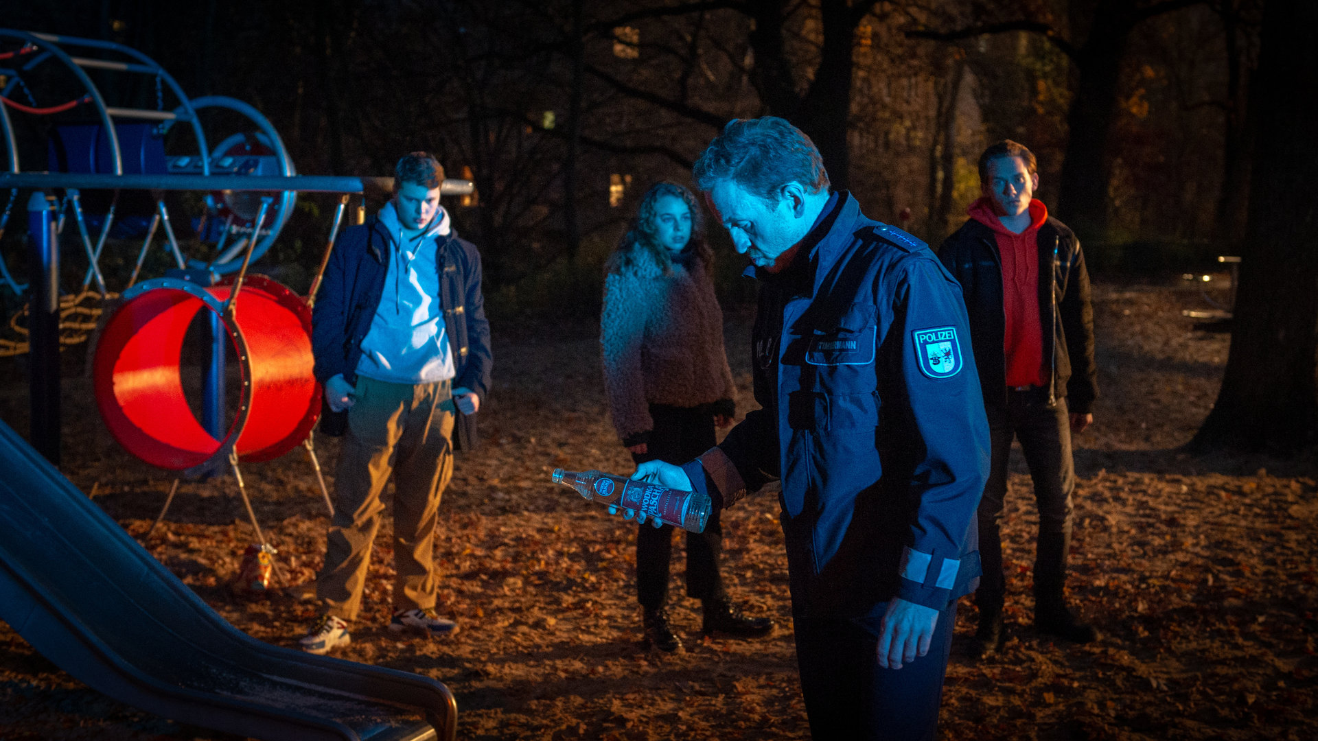 "SOKO Wismar - Am falschen Ort": Es ist Nacht. Ein Spielplatz ist in fahles Licht getaucht. Timmermann (Mathias Junge) hält eine Schnapsflasche in der Hand, die er Simon Hofmeister (Elmo Anton Stratz) abgenommen hat. Bei Simon stehen seine Freunde Violetta (Amelie Herres) und Marek (Alvar Goetze). Simon blickt betreten zu Boden. Violetta und Marek wirken angespannt.
