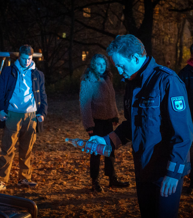 "SOKO Wismar - Am falschen Ort": Es ist Nacht. Ein Spielplatz ist in fahles Licht getaucht. Timmermann (Mathias Junge) hält eine Schnapsflasche in der Hand, die er Simon Hofmeister (Elmo Anton Stratz) abgenommen hat. Bei Simon stehen seine Freunde Violetta (Amelie Herres) und Marek (Alvar Goetze). Simon blickt betreten zu Boden. Violetta und Marek wirken angespannt.