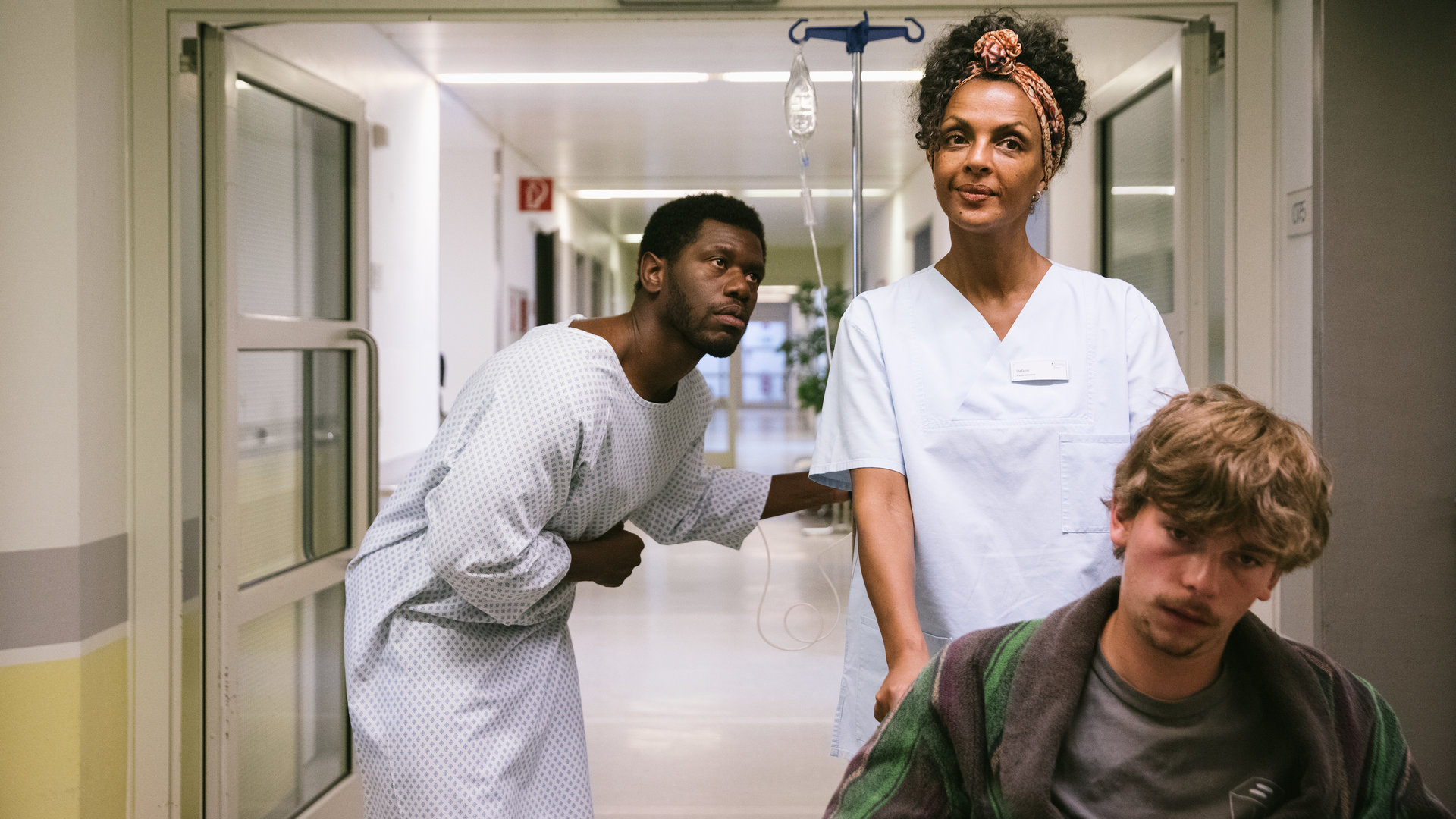 "I don't work here - Friedhof": Dawit (Akeem van Flodrop) steht gebückt in einem Krankenhausflur und hält sich mit der rechten Hand den Bauch. Er trägt ein weißes Krankenhaushemd und hält sich mit links an einem Infusionsständer fest. Er schaut seiner Mutter Lemlem (Dennenesch Zoudé) hinterher, die in ihrer Pflegerinnenkleidung einen Patienten im Rollstuhl vor sich herschiebt und dabei lächelt.