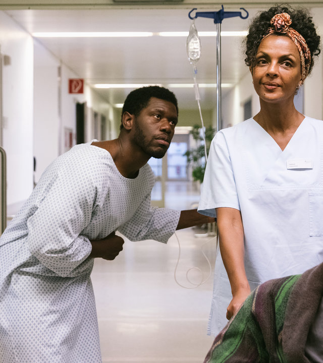"I don't work here - Friedhof": Dawit (Akeem van Flodrop) steht gebückt in einem Krankenhausflur und hält sich mit der rechten Hand den Bauch. Er trägt ein weißes Krankenhaushemd und hält sich mit links an einem Infusionsständer fest. Er schaut seiner Mutter Lemlem (Dennenesch Zoudé) hinterher, die in ihrer Pflegerinnenkleidung einen Patienten im Rollstuhl vor sich herschiebt und dabei lächelt.