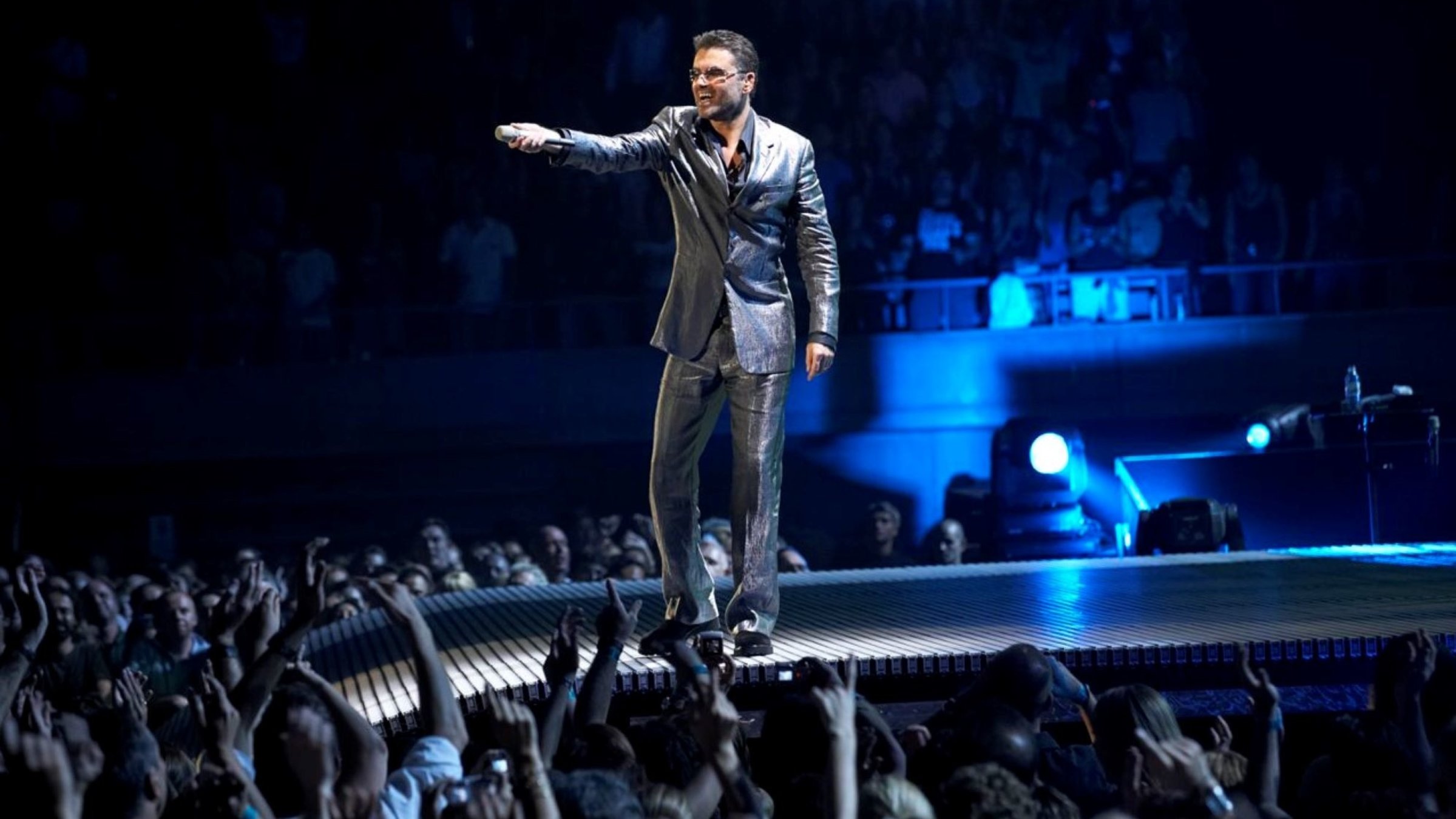 "George Michael:  Live in London - 25 Live Tour": Der Sänger George Michael steht auf einem Steg der von einer Bühne ins Publikum führt und hält ein Mikrofon in Richtung der Zuschauer, die zum Teil ihre Arme in die Höhe strecken. Bekleidet ist der Sänger mit einem silberfarbenen Anzug.