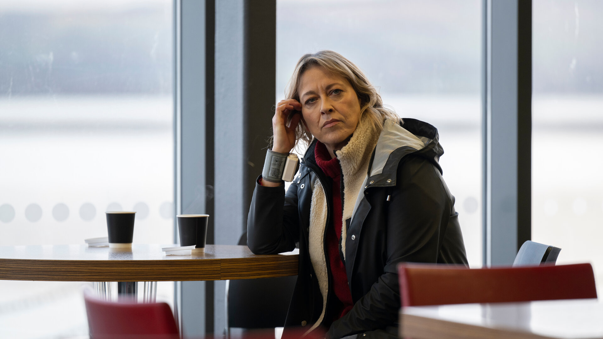 "Annika – Mord an Schottlands Küste - Wahre Werte": Annika (Nicola Walker) sitzt vor einer Fensterfront in einer Cafeteria an einem Tisch, auf dem zwei Becher stehen. An ihrem rechten Handgelenk trägt sie ein Pulsmessgerät.