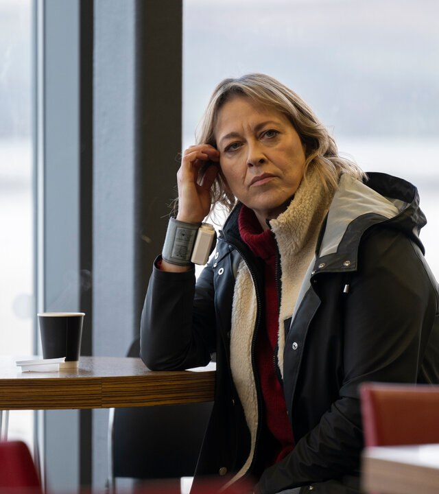 "Annika – Mord an Schottlands Küste - Wahre Werte": Annika (Nicola Walker) sitzt vor einer Fensterfront in einer Cafeteria an einem Tisch, auf dem zwei Becher stehen. An ihrem rechten Handgelenk trägt sie ein Pulsmessgerät.