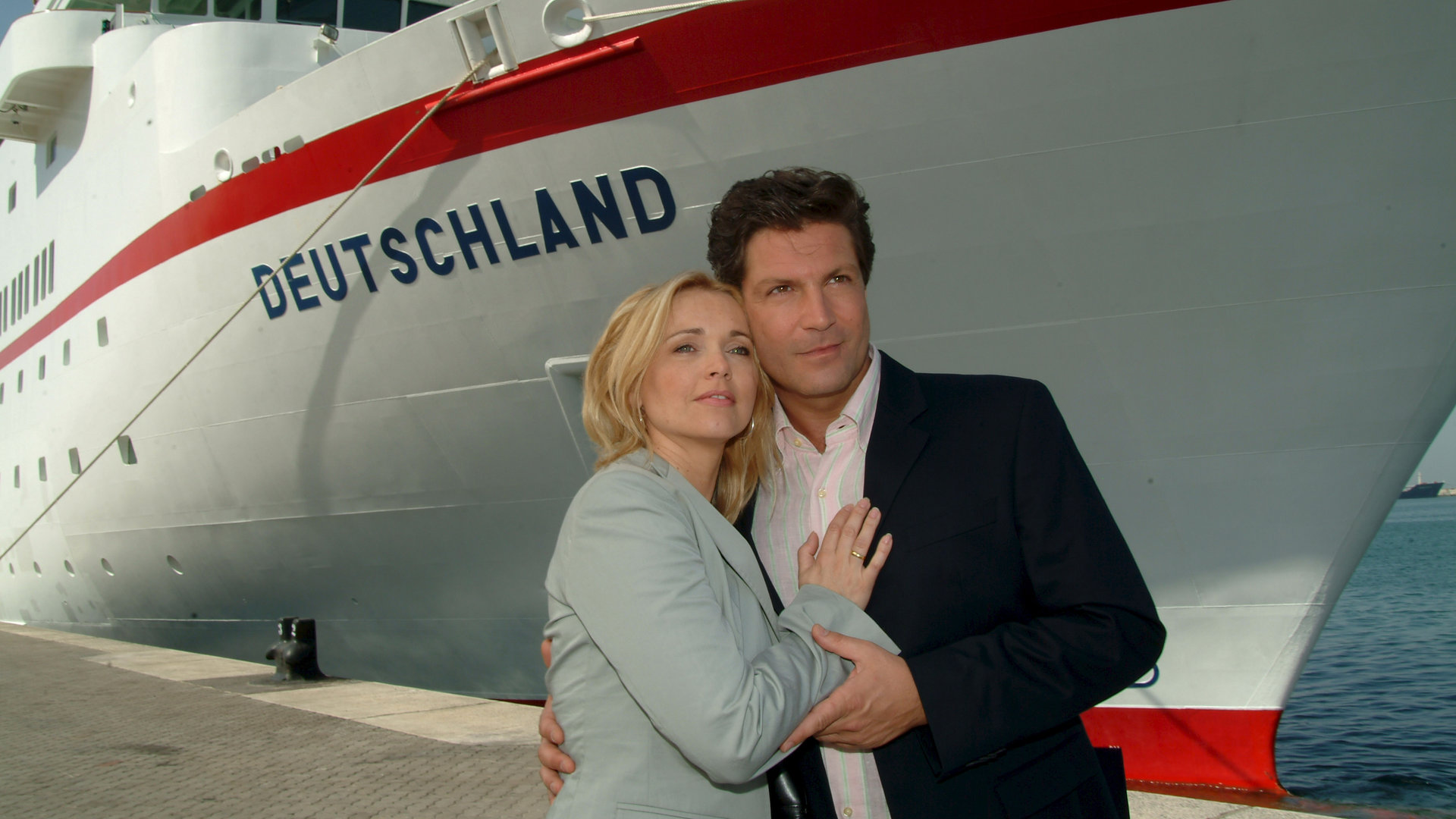 "Das Traumschiff - Shanghai": Meret (Tina Ruland) und Paul (Francis Fulton-Smith) stehen Arm in Arm vor der MS Deutschland, die im Hafen liegt. Beide schauen verträumt in die Ferne.