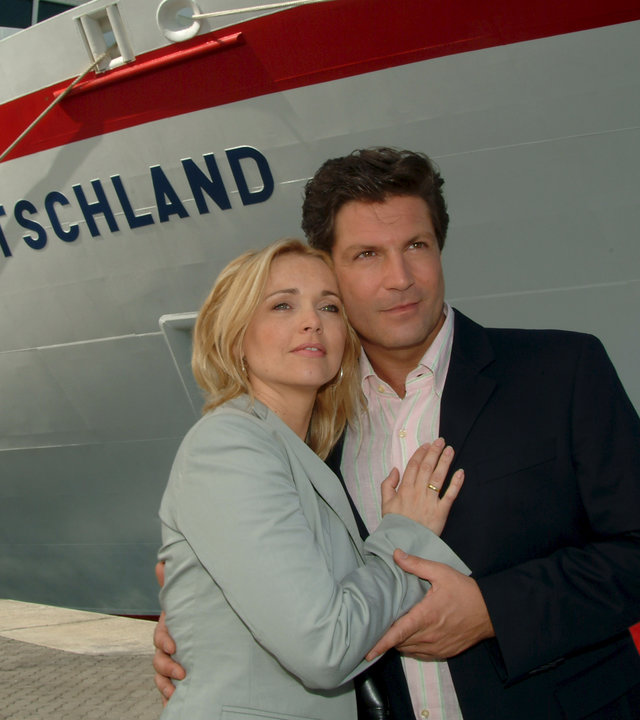"Das Traumschiff - Shanghai": Meret (Tina Ruland) und Paul (Francis Fulton-Smith) stehen Arm in Arm vor der MS Deutschland, die im Hafen liegt. Beide schauen verträumt in die Ferne.