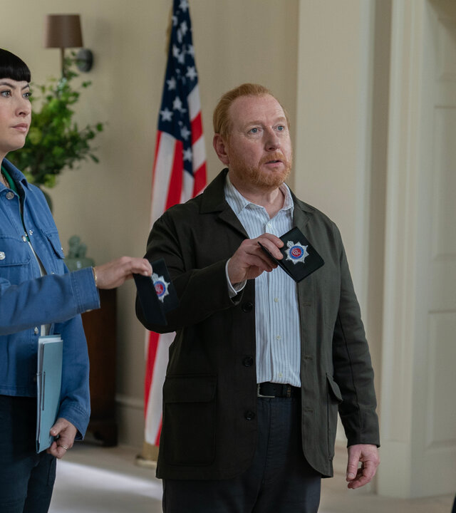 "The Chelsea Detective - Tod im Kleingarten": Die leger gekleideten Max Arnold (Adrian Scarborough) und Layla Walsh (Vanessa Emme) stehen in einem elegant ausgestatteten Flur und zeigen dem ihnen gegenüberstehenden Nick (Greg Lockett) ihre Dienstausweise des Chelsea CID. Nick trägt einen dunkelblauen Anzug. Im Hintergrund ist eine US-amerikanische Flagge zu sehen.