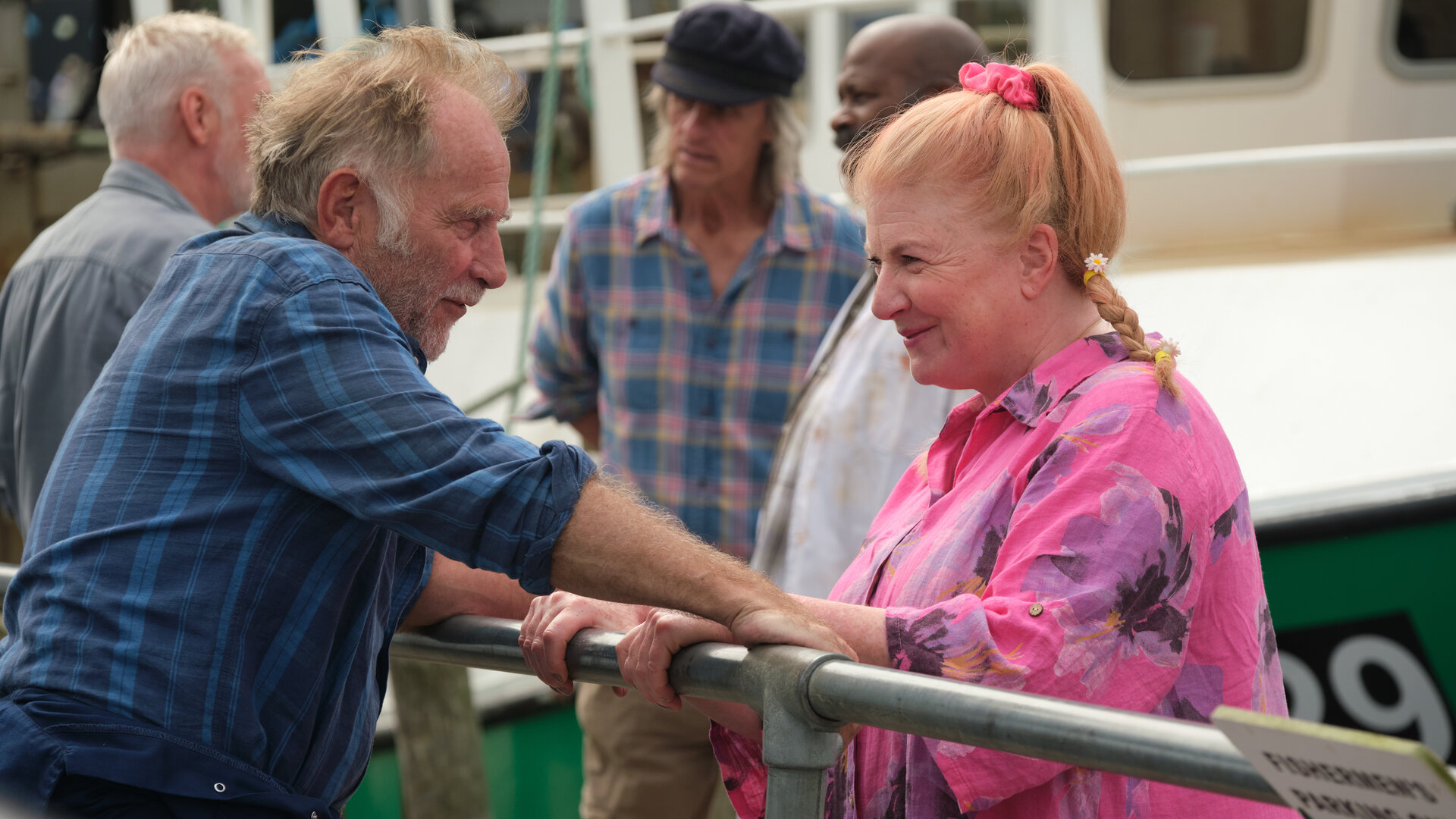 "Beyond Paradise - Über Bord": Jaimie (Danny Webb) und Margo Martins (Felicity Montagu) stehen sich an einem Geländer gegenüber und schauen sich lächelnd an. Im Hintergrund unterhalten sich drei Männer (Komparsen) vor einem Fischerboot.