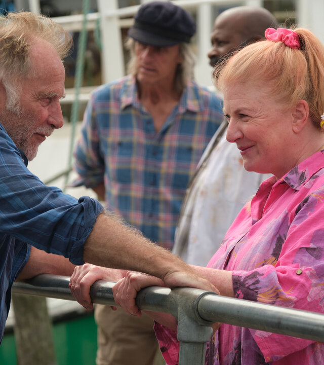 "Beyond Paradise - Über Bord": Jaimie (Danny Webb) und Margo Martins (Felicity Montagu) stehen sich an einem Geländer gegenüber und schauen sich lächelnd an. Im Hintergrund unterhalten sich drei Männer (Komparsen) vor einem Fischerboot.