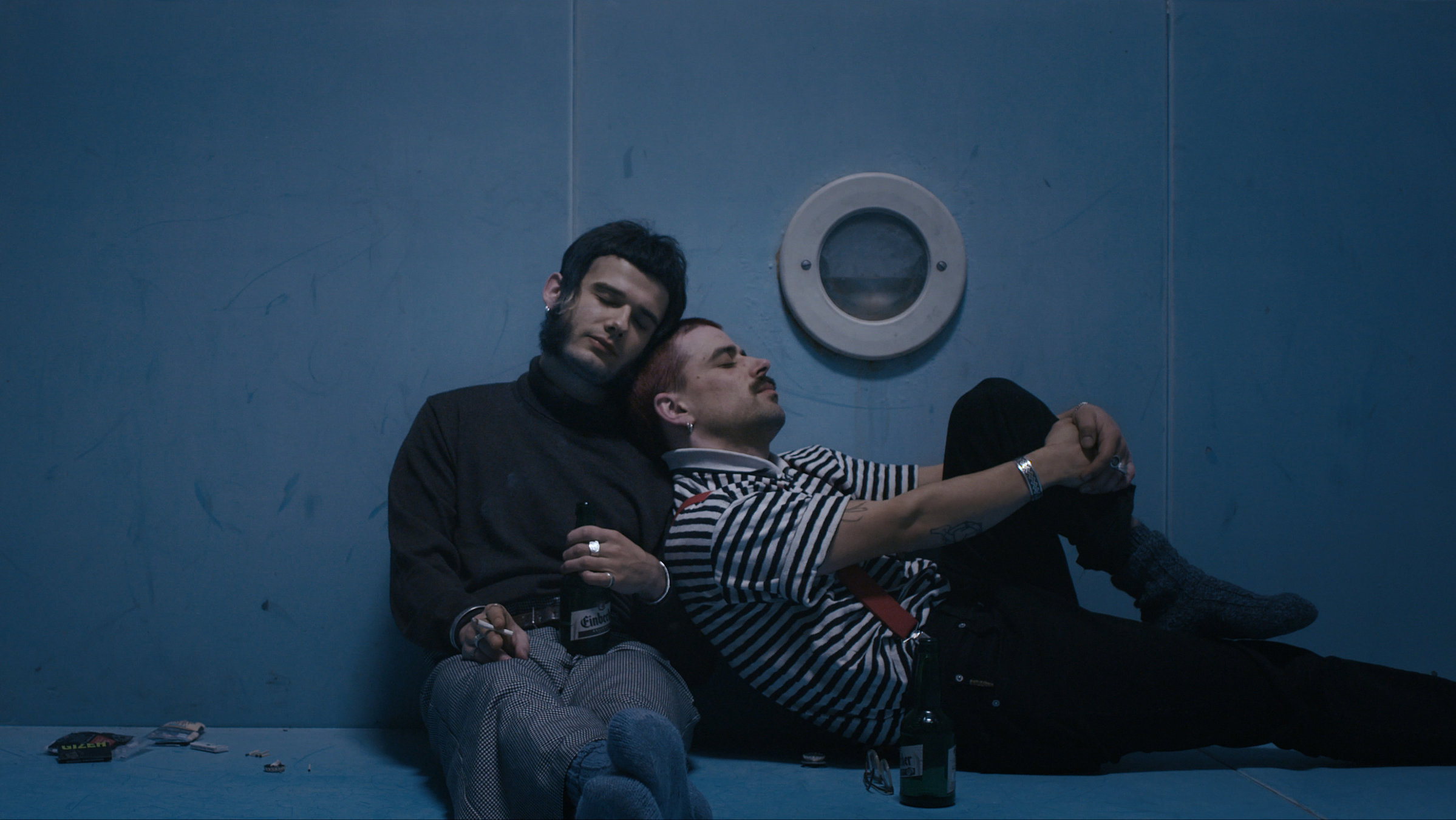 "Ab 18! – Fehler und Irritation": Zwei junge Männer sitzen in dämmrigem Licht auf dem Boden eines Swimming Pools, in dem kein Wasser ist. Der links sitzende mit Rollkragenpullover lehnt an die Mauer des Pools. Er hält eine brennende Zigarette in der – vom Betrachter aus gesehen – linken und eine Bierflasche in der rechten Hand. An ihn lehnt sich der zweite Mann mit Schnurrbart und blau gestreiftem T-Shirt, der im 90°-Winkel zu ihm sitzt. Er hat ein Bein angezogen. Beide haben die Augen geschlossen.
