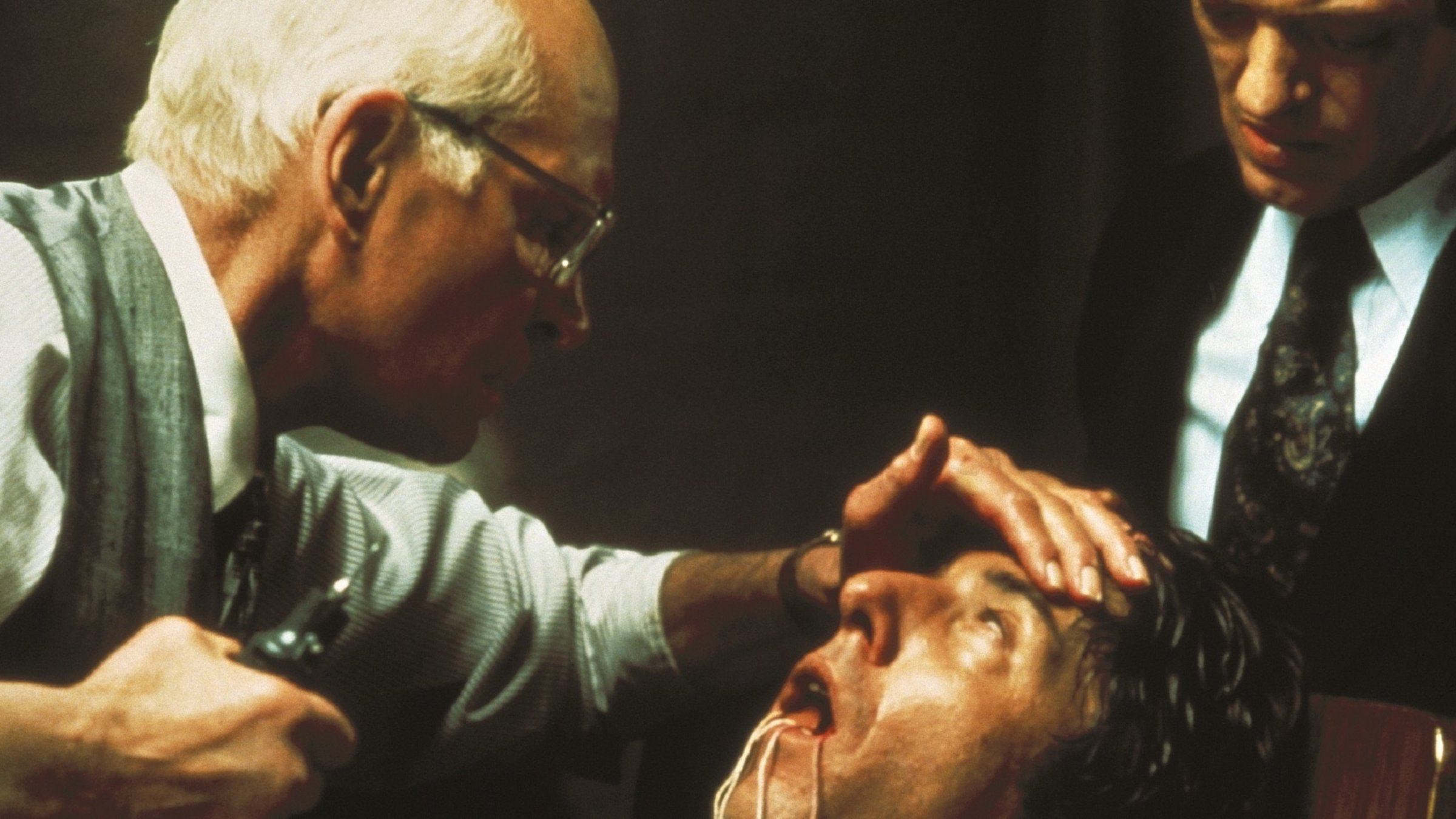 "Der Marathon-Mann": Ein älterer Mann (Sir Laurence Olivier) mit Halbglatze, Brille, fein gekleidet in Hemd, Weste und Krawatte, in der Hand einen Zahnarzt-Bohrer, beugt sich von links über einen halb bewusstlosen, ihm gegenüber sitzenden Mann, aus dessen offenem Mund Fäden hängen. Am rechten Bildrand steht ein weiterer Mann in Anzug und Krawatte, der die Situation beobachtet.