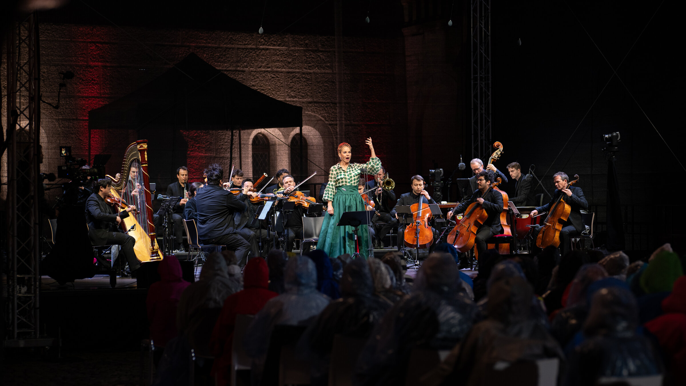 "Neuschwanstein Konzerte 2025: Elīna Garanča": Das Bild zeigt eine Bühne, auf der im Mittelpunkt die Sängerin in einem grünen Rock und einer grün gepunkteten Bluse steht. Sie hat den linken Arm gebeugt und in die Höhe gestreckt. Um sie herum sitzen die Orchestermusiker*innen, ebenfalls in festlicher Kleidung. Die Bühne ist in leichten roten und blauen Tönen beleuchtet.