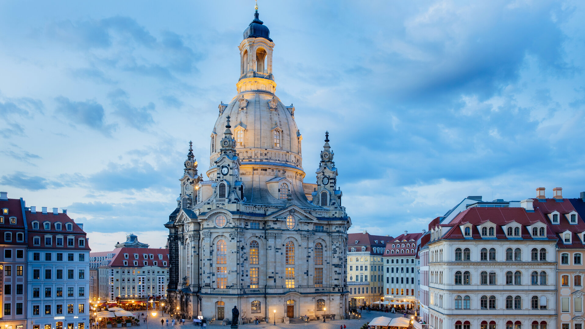 "Festlicher Neujahrsgottesdienst - Siehe, ich mache alles neu!": Inmitten der restaurierten Altstadt steht die Dresdner Frauenkirche. Sie ist umgeben von Häusern, im Vordergrund befindet sich der Neumarkt. In der beginnenden Abenddämmerung sind die Frauenkirche und der Marktplatz beleuchtet.