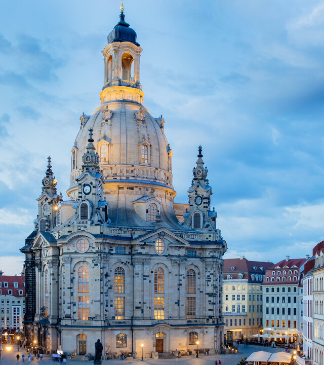 "Festlicher Neujahrsgottesdienst - Siehe, ich mache alles neu!": Inmitten der restaurierten Altstadt steht die Dresdner Frauenkirche. Sie ist umgeben von Häusern, im Vordergrund befindet sich der Neumarkt. In der beginnenden Abenddämmerung sind die Frauenkirche und der Marktplatz beleuchtet.