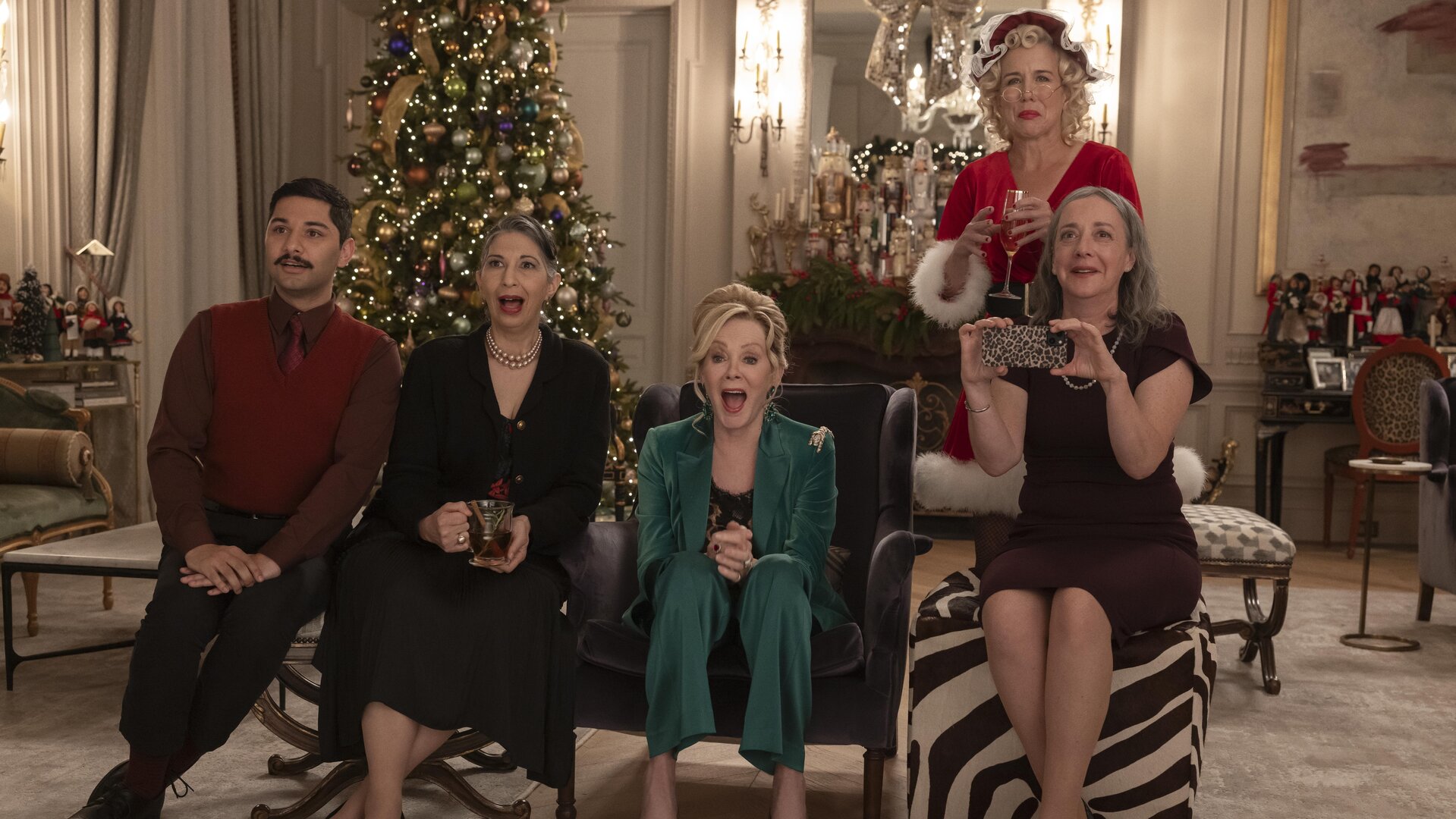 "Hacks - Das Vegas-Weihnachtsspektakel": In festlicher Kleidung sitzen Damien (Mark Indelicato), Josefina (Rose Abdoo), Deborah (Jean Smart) und Avas Mutter Nina (Jane Adams) lachend nebeneinander in einem festlich geschmückten Wohnraum. Hinter ihnen steht Bürgermeisterin Mayor Jo (Lauren Weedman) im Santa-Kostüm. An der Seite steht ein großer Weihnachtsbaum.