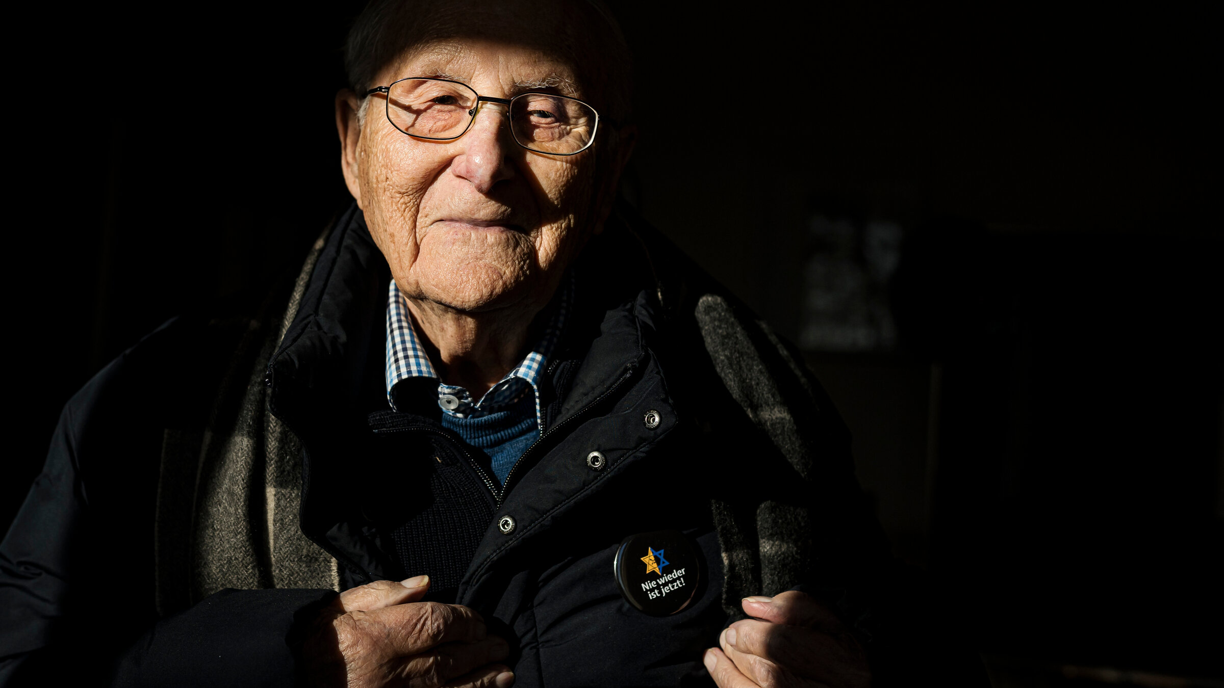 "37°: Die Nummer auf meinem Arm - Albrecht Weinberg und seine Geschichte": Der 99-jährige Holocaust-Überlebende Albrecht Weinberg steht in seinem Zimmer in Leer, Ostfriesland. Er ist bereits mit dicker Winterkleidung zum Rausgehen angezogen. Ein Sonnenstrahl fällt durchs Fenster auf Teile seines Gesichts und Oberkörpers. Das Zimmer hinter ihm ist im Schatten und kaum zu sehen. An seiner Brust trägt er einen Button mit der Aufschrift "Nie wieder ist jetzt" und einem blau-gelben Davidstern.
