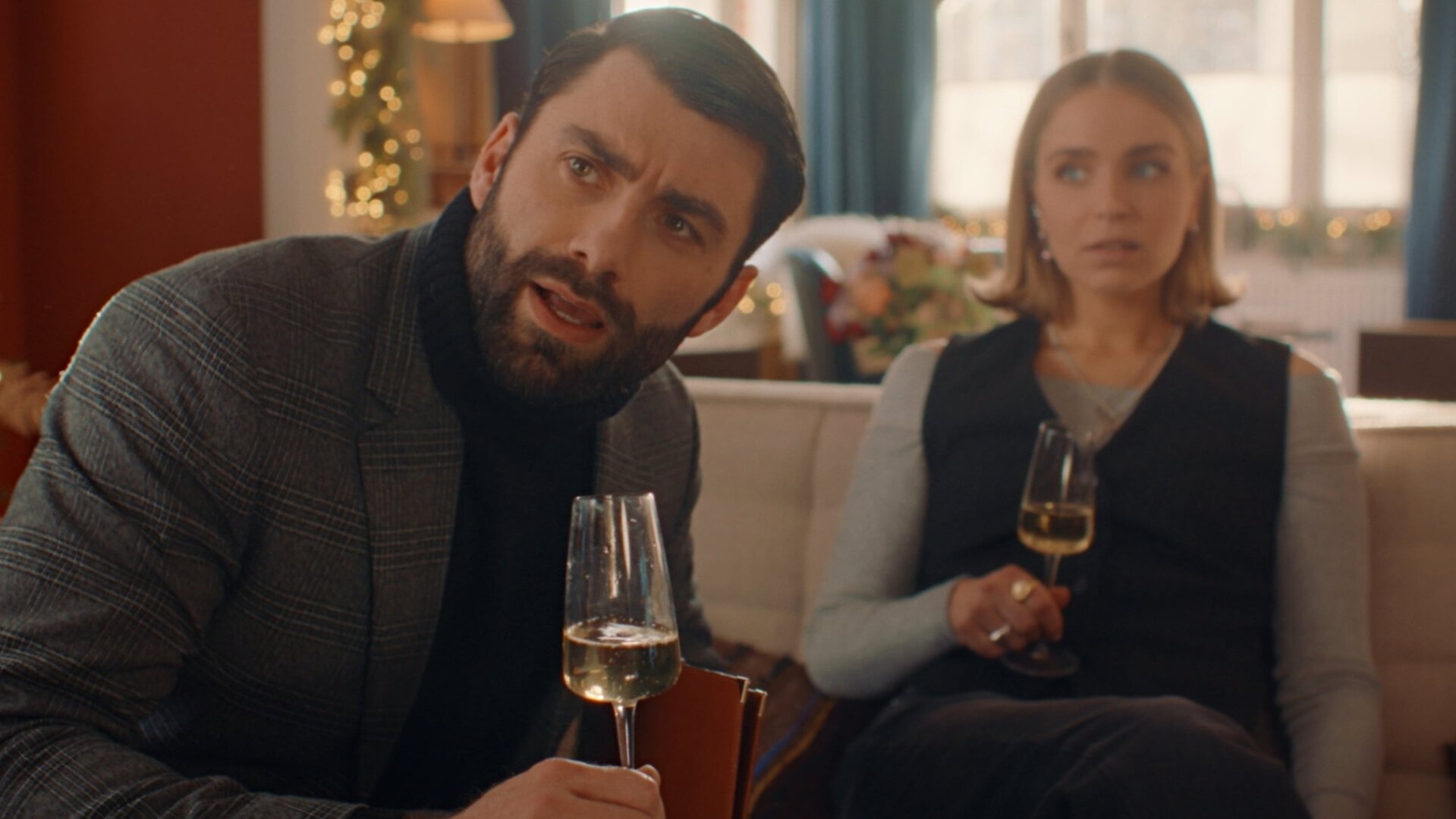 "Single Bells - 12 Dates till Christmas: Laurent": Laurent (Sieg De Doncker) und Mina (Julie Van den Steen) sitzen mit einem Glas Weißwein in der Hand auf Sofas im Wohnzimmer. Laurent schaut prüfend zur Seite, während ihn Mina im Hintergrund irritiert anschaut.