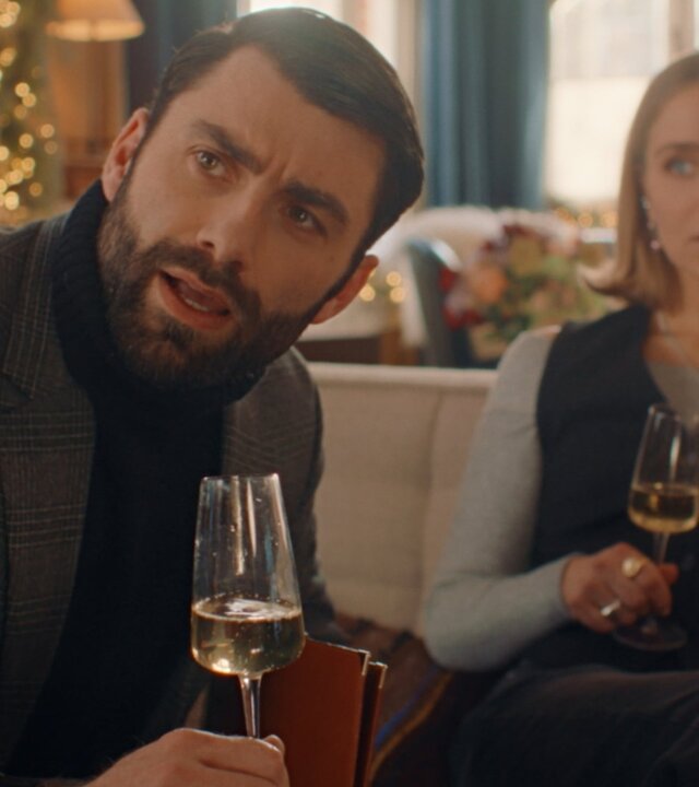 "Single Bells - 12 Dates till Christmas: Laurent": Laurent (Sieg De Doncker) und Mina (Julie Van den Steen) sitzen mit einem Glas Weißwein in der Hand auf Sofas im Wohnzimmer. Laurent schaut prüfend zur Seite, während ihn Mina im Hintergrund irritiert anschaut.