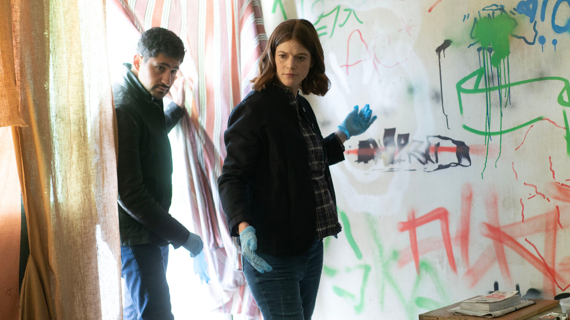 "Vigil - Tödliche Drohnen: Wüstenstaat": Kirsten Longacre (Rose Leslie) und ihr Kollege Daniel Ramsay (Amir El-Masry) stehen in einer Wohnung vor einen bekritzelten Wand und schauen sich um.